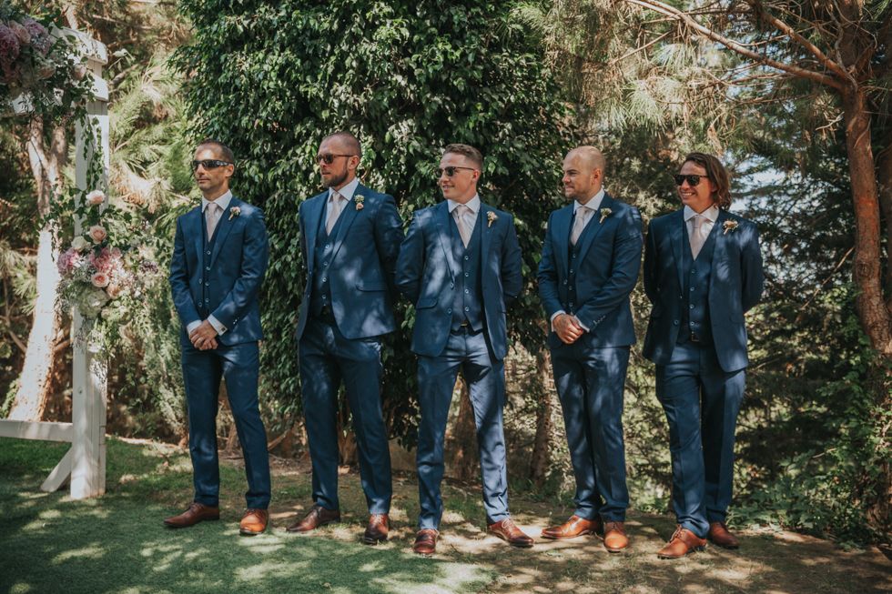 Five groomsmen in matching navy suits and brown dress shoes stand in line on garden path.