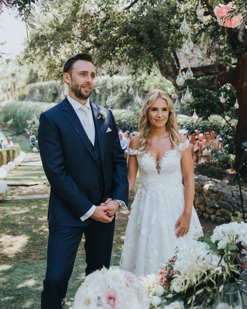 A couple in wedding attire poses together in a garden setting with white and pink floral arrangements.