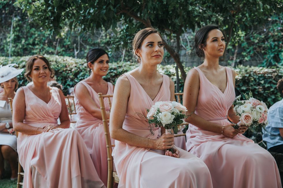 Bridesmaids in flowing pink dresses sit together holding white bouquets during outdoor wedding ceremony.
