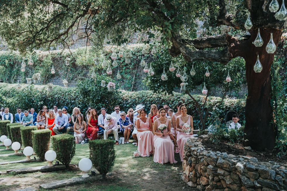 Wedding guests in colorful attire seated outdoors under trees during an elegant garden ceremony on a sunny day.