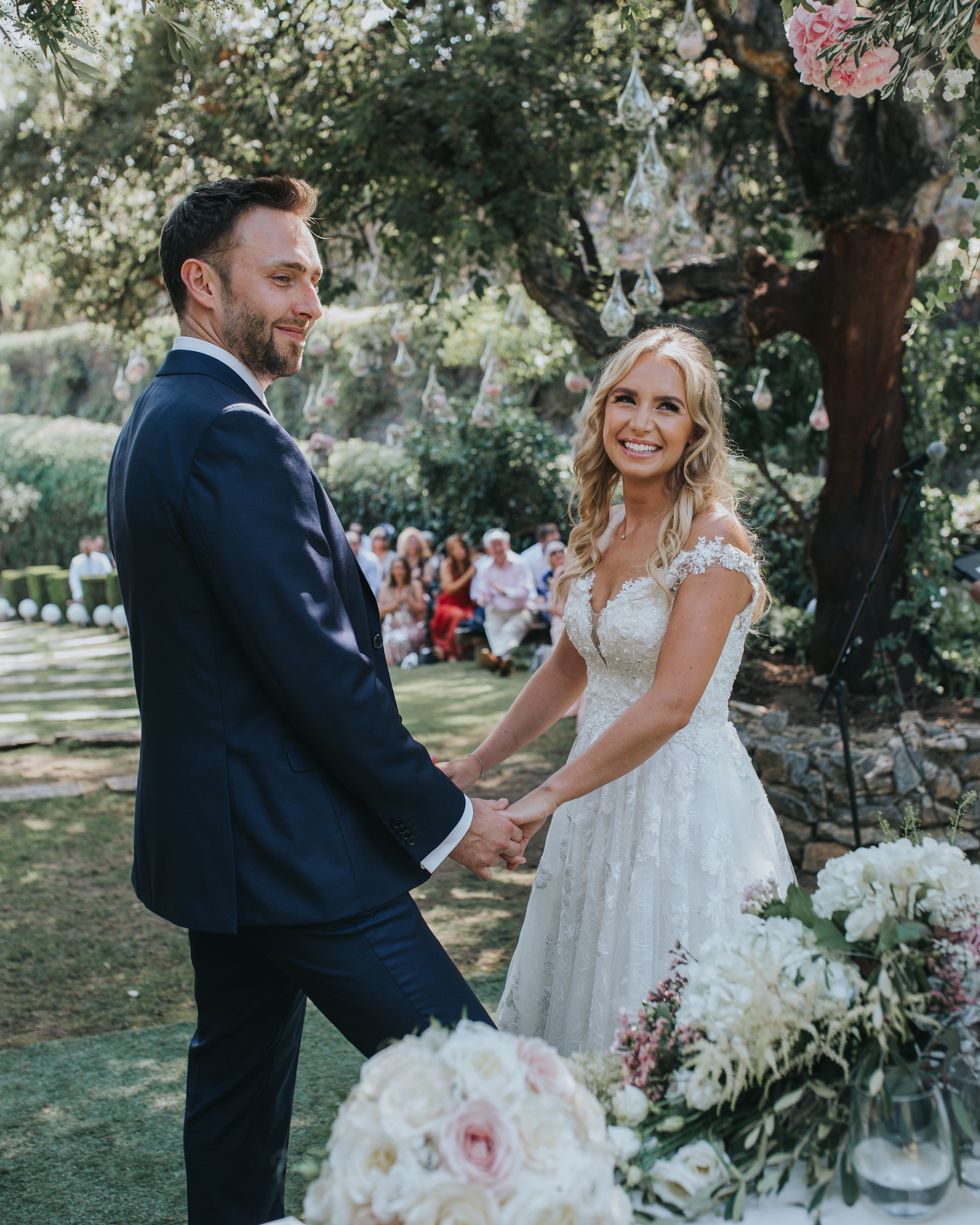 A bride and groom hold hands in front of wedding guests at an outdoor garden ceremony with floral arrangements.