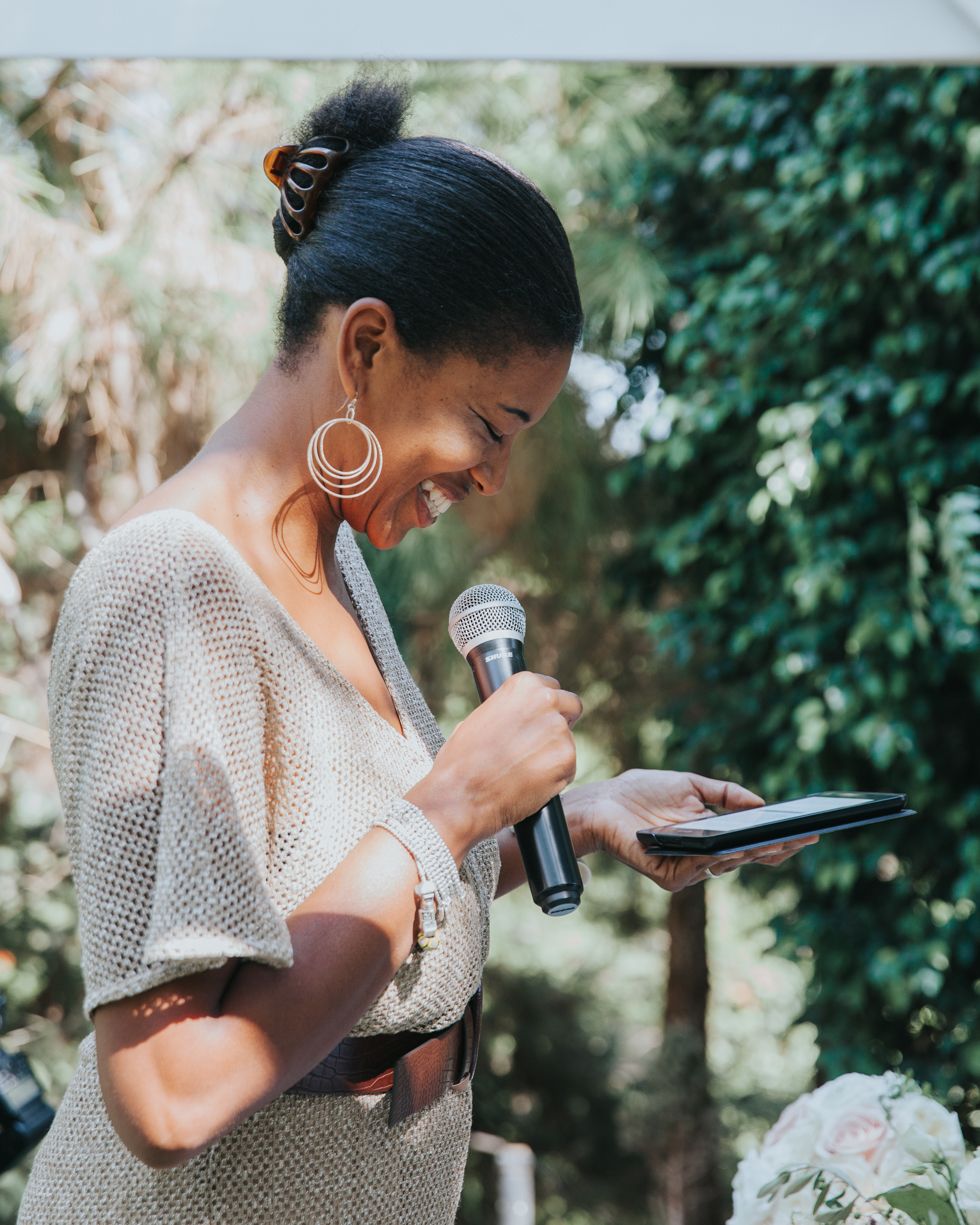 A wedding speaker in a beige dress holding a microphone and looking down at notes during an outdoor ceremony.