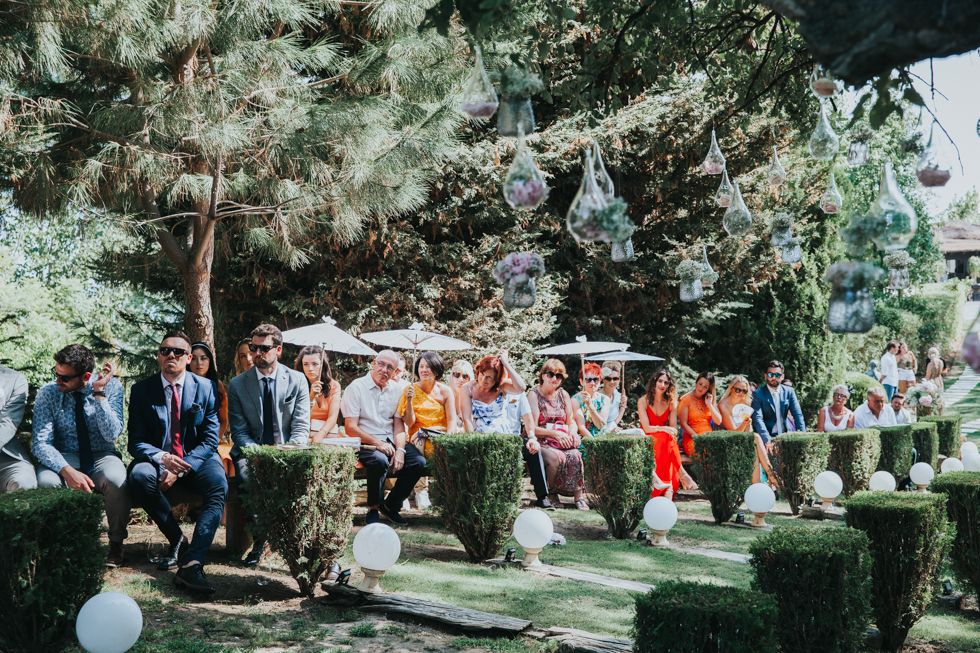 A group of wedding guests sit in rows of chairs outdoors under shaded trees during a sunny garden ceremony.