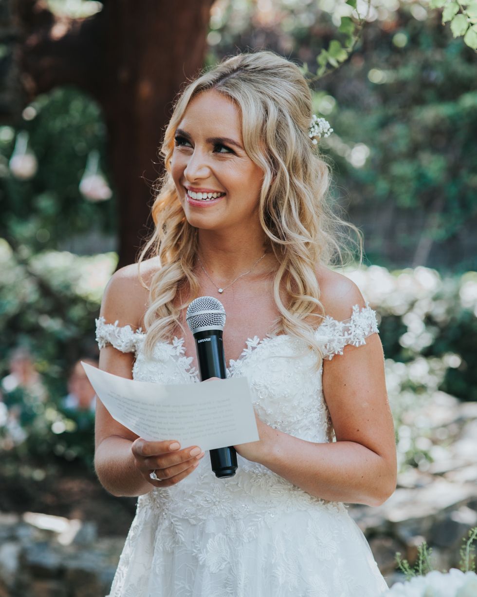 A bride in a white lace dress giving a speech while holding a microphone and notes at an outdoor wedding.