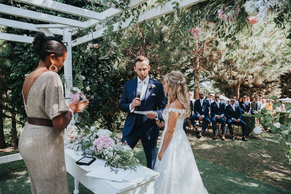 Garden wedding ceremony under a white pergola with floral decorations and guests seated on lawn in summertime.