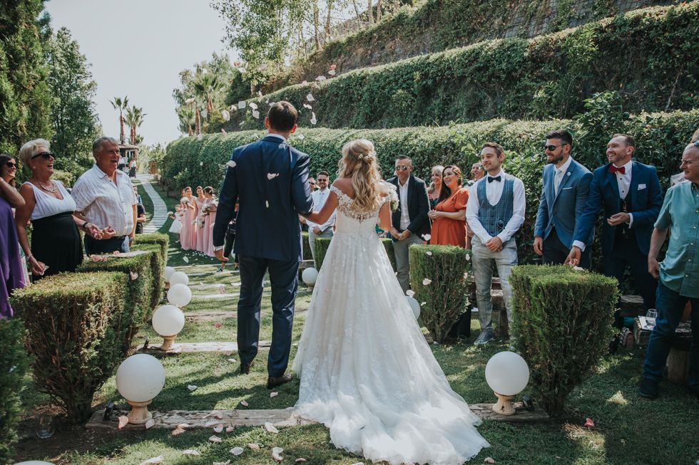 Outdoor garden wedding ceremony with couple standing between manicured hedges and white balloons along the aisle.