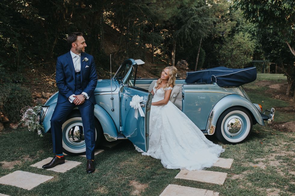 A bride and groom pose with a vintage blue convertible car in a rustic outdoor wedding setting.
