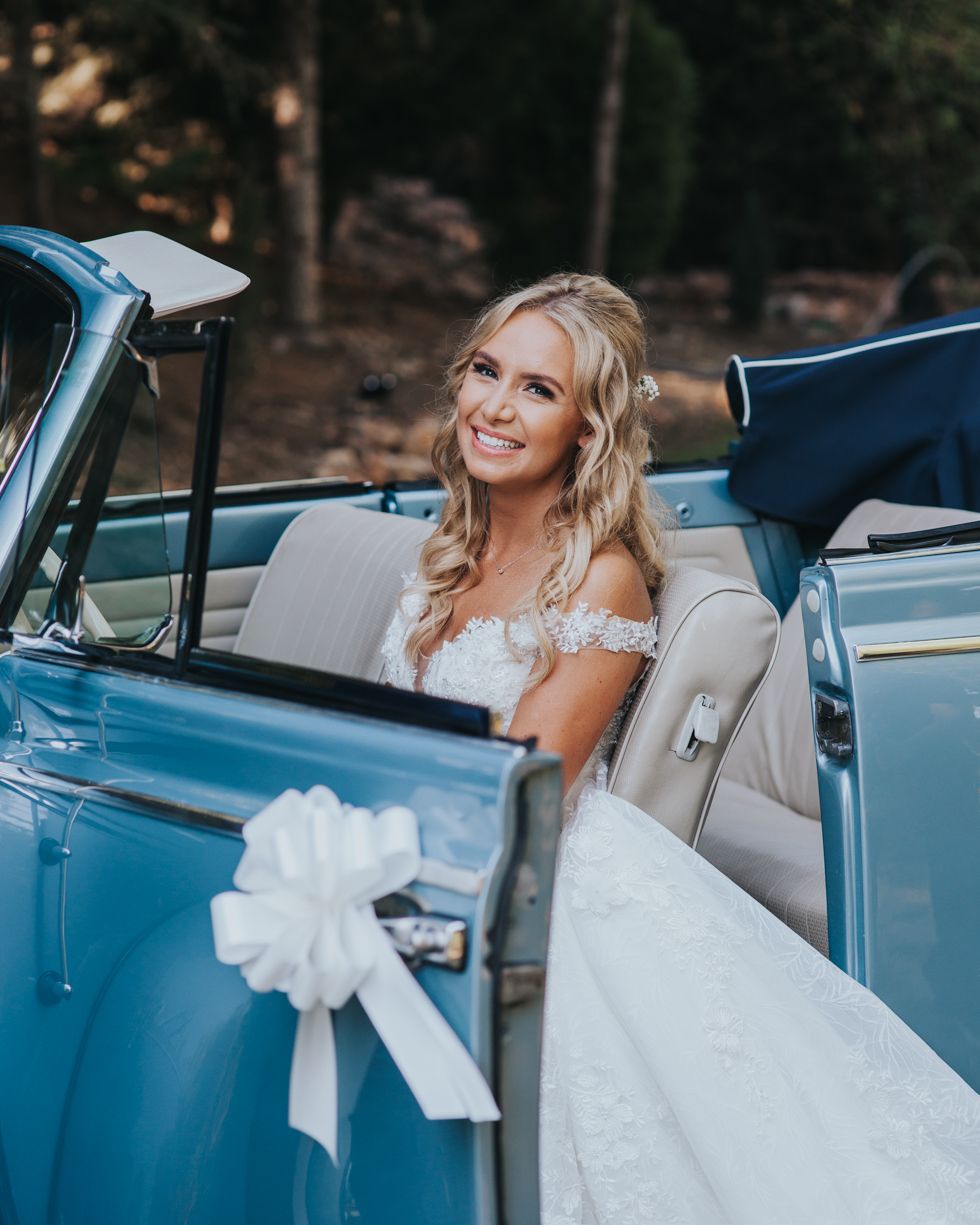 A person in a white lace wedding dress sits in a vintage blue convertible car decorated with a white bow.