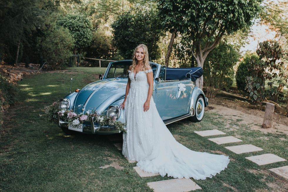 A classic blue Volkswagen Beetle convertible parked on a garden path serves as backdrop for an elegant wedding photo.
