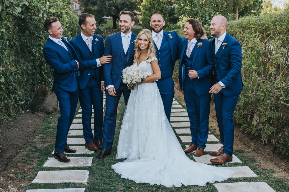 Wedding party in navy blue suits stands with bride on stone garden path surrounded by greenery.