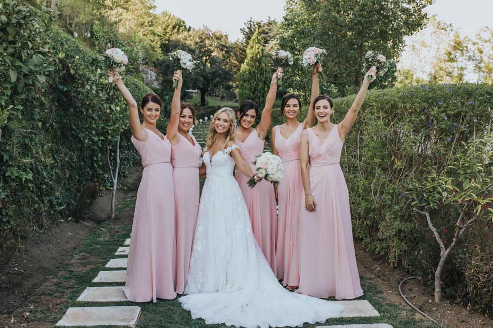 A bride in white gown poses with bridesmaids wearing blush pink dresses holding flower bouquets on a garden path.