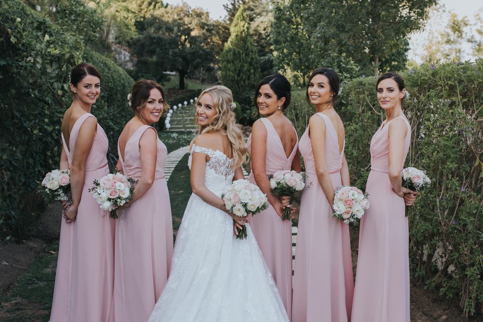 Wedding party poses outdoors in matching blush pink bridesmaid dresses and white bridal gown holding floral bouquets.
