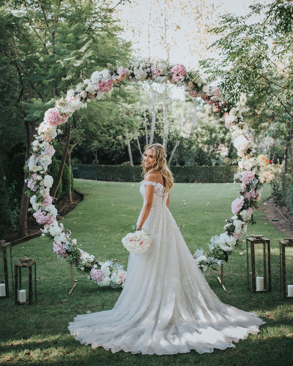 Elegant wedding gown with flowing train under a romantic floral arch with pink and white flowers in a garden setting.