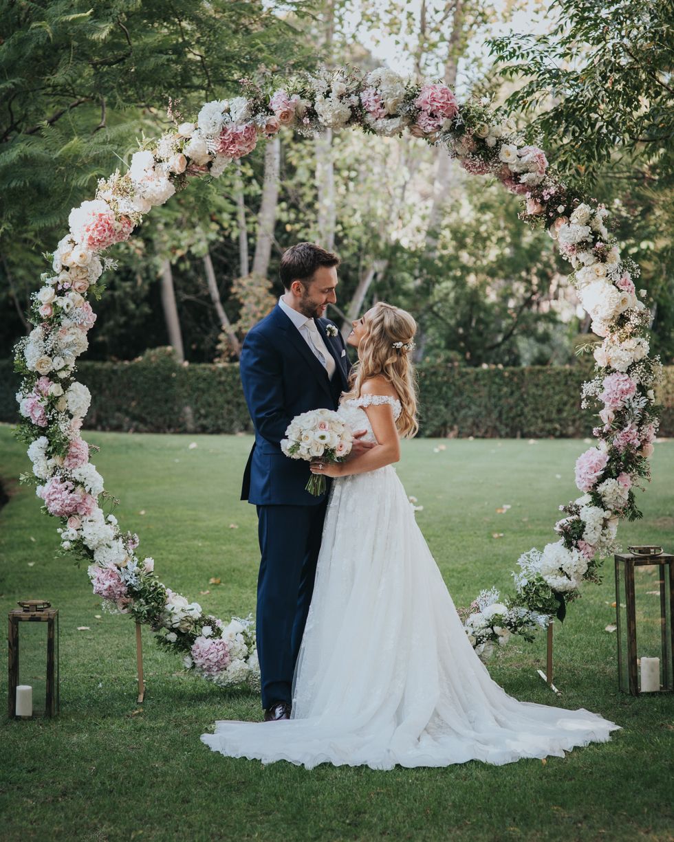 A romantic wedding ceremony moment under a curved arch adorned with pink and white blooms in a garden setting.