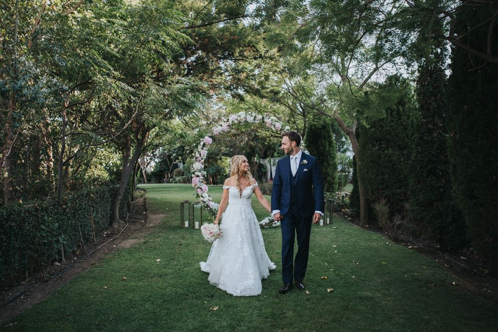 A couple in wedding attire walks hand in hand through a garden archway covered in white flowering vines.
