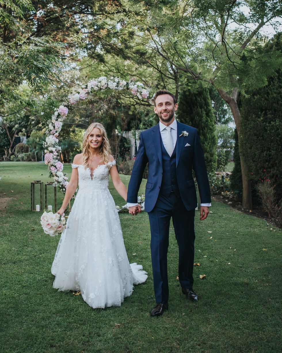 A wedding couple holds hands under a floral arch in a lush garden setting with trees and green lawn.