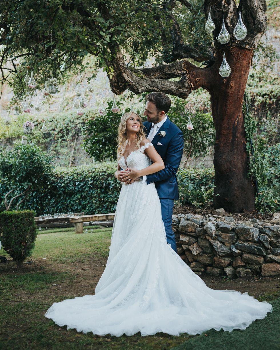 A couple shares a romantic moment in a garden setting with hanging glass decorations and stone walls in the background.
