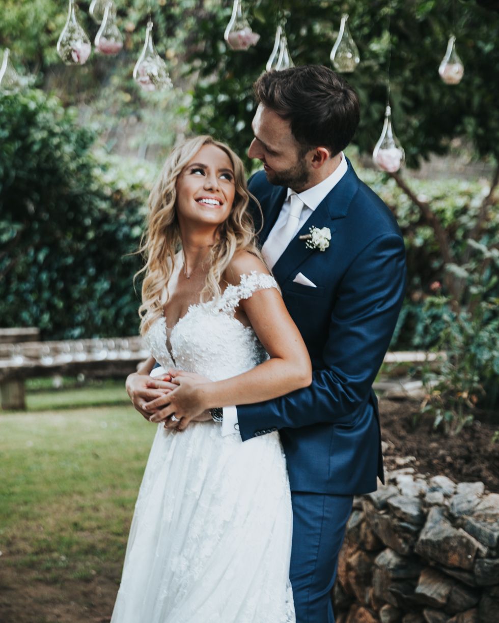 Romantic outdoor wedding photo with hanging glass bulb decorations and couple embracing on a lawn with greenery background.
