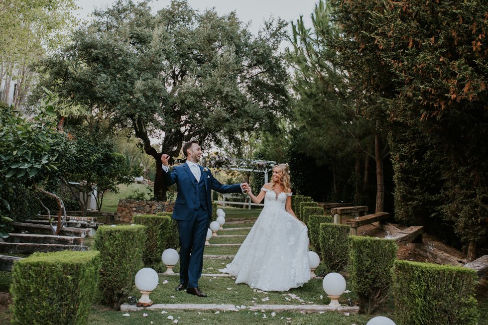Wedding couple stands together on garden steps surrounded by greenery and illuminated round lanterns.