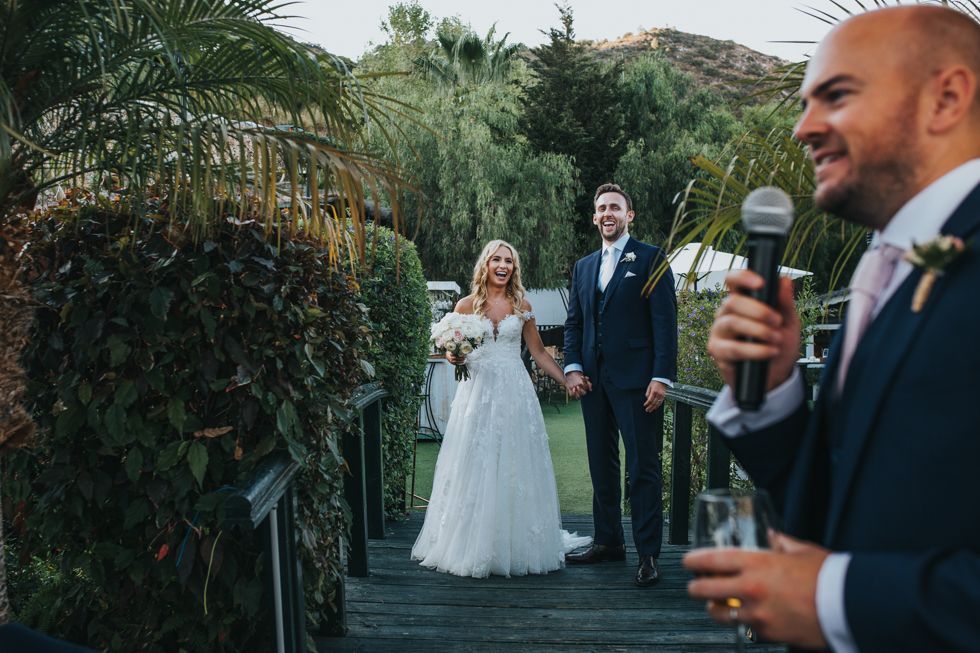 Wedding ceremony captured at outdoor garden venue with speaker giving speech on wooden deck surrounded by lush greenery.