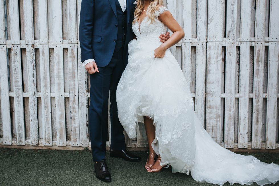 A bride wearing a white lace wedding gown and casual flip flops stands next to groom in navy suit against white fence.