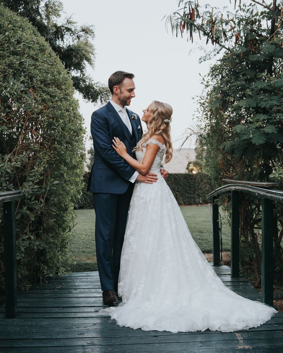 A couple embraces on a wooden bridge surrounded by lush greenery in an elegant wedding photoshoot.