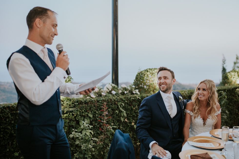 Best man delivers wedding speech outdoors during reception at scenic mountain venue with greenery and guests seated nearby.