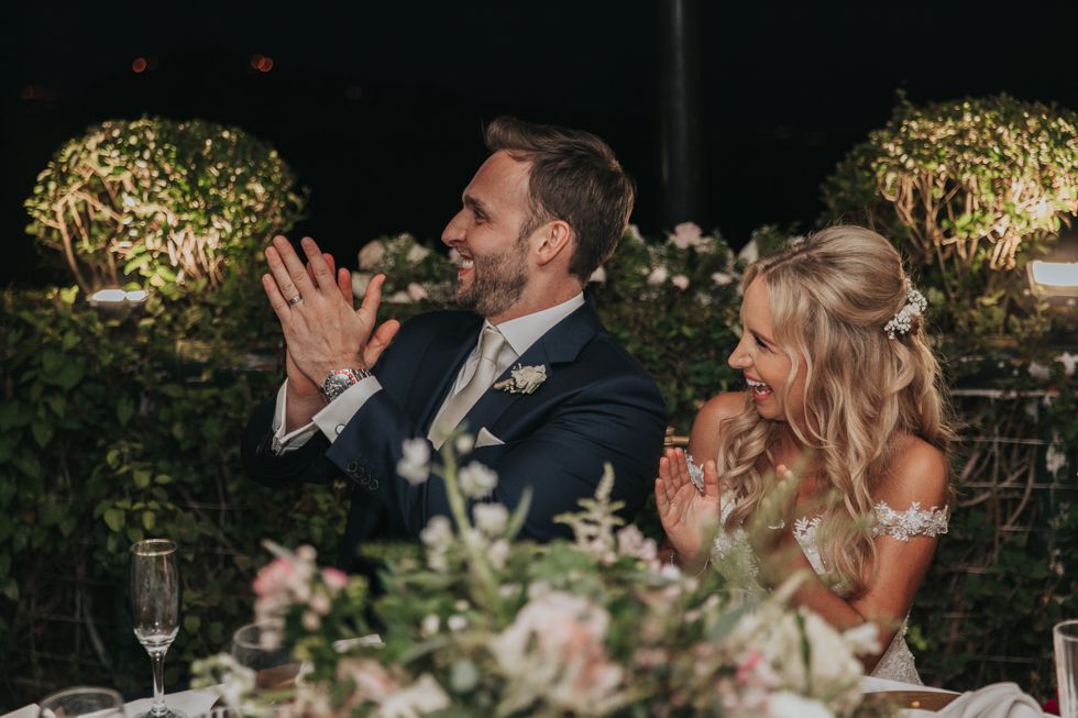 Newlyweds share joyful moment at reception dinner surrounded by floral arrangements and twinkling lights.