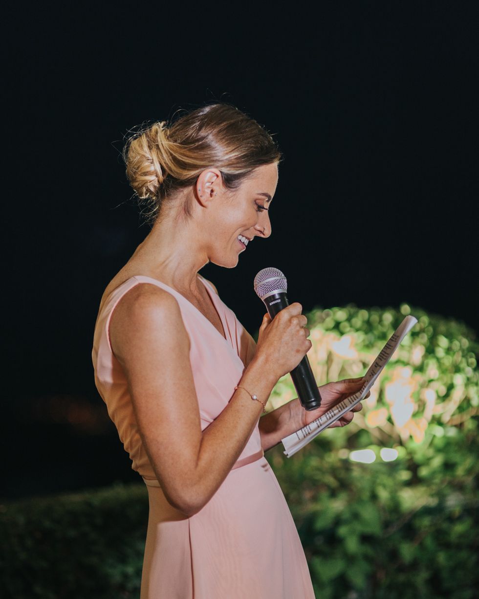 Speaking into a microphone in an elegant pink dress, a woman smiles while giving a speech outdoors.