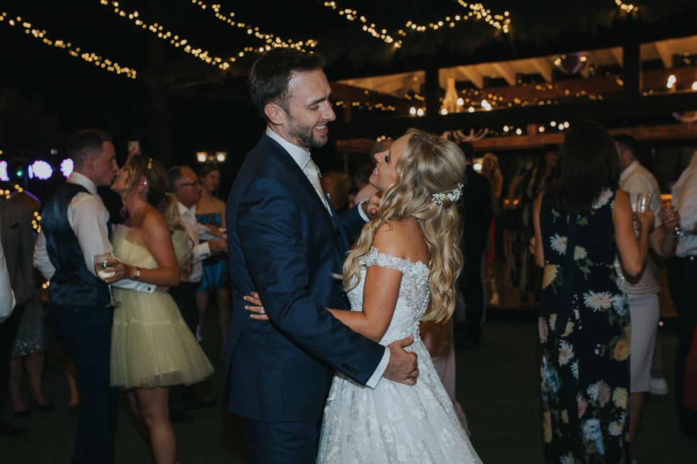 A couple shares a romantic dance at a wedding reception under twinkling lights in a rustic indoor venue.