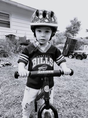 A small child rides a black balance bike while wearing a Bills jersey and helmet.