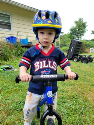 A toddler in a Bills jersey and blue helmet stands with a balance bike in the grass.