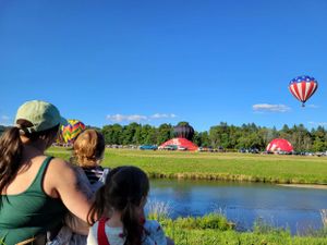 Hot air balloons float over a scenic lake landscape on a bright summer day.