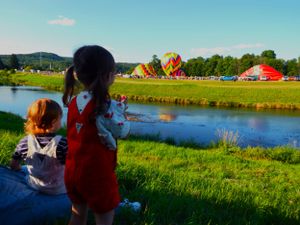 Hot air balloons floating over a grassy field near a lake during a summer festival.
