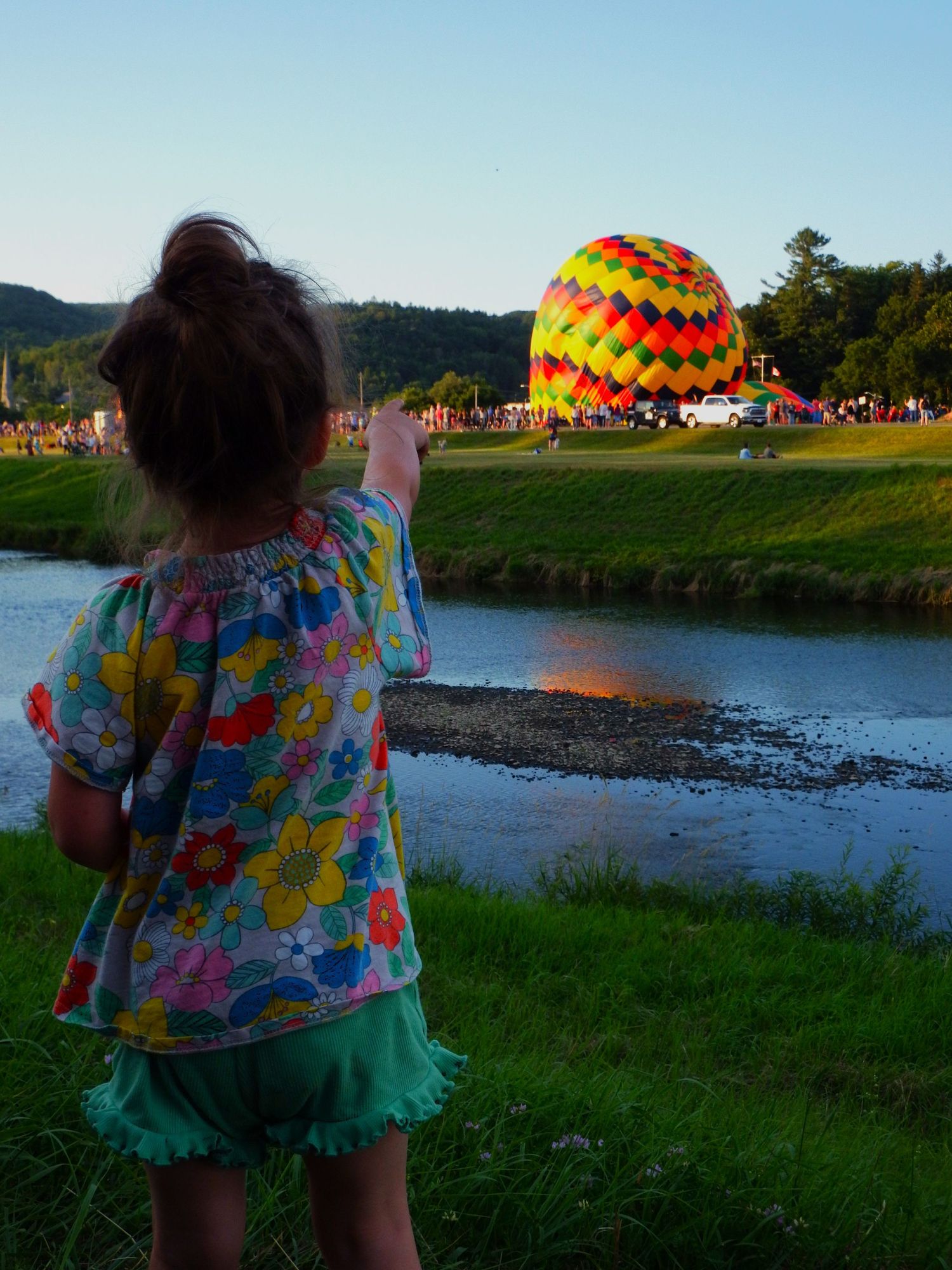Person in floral dress watching hot air balloons by a lake during sunset.