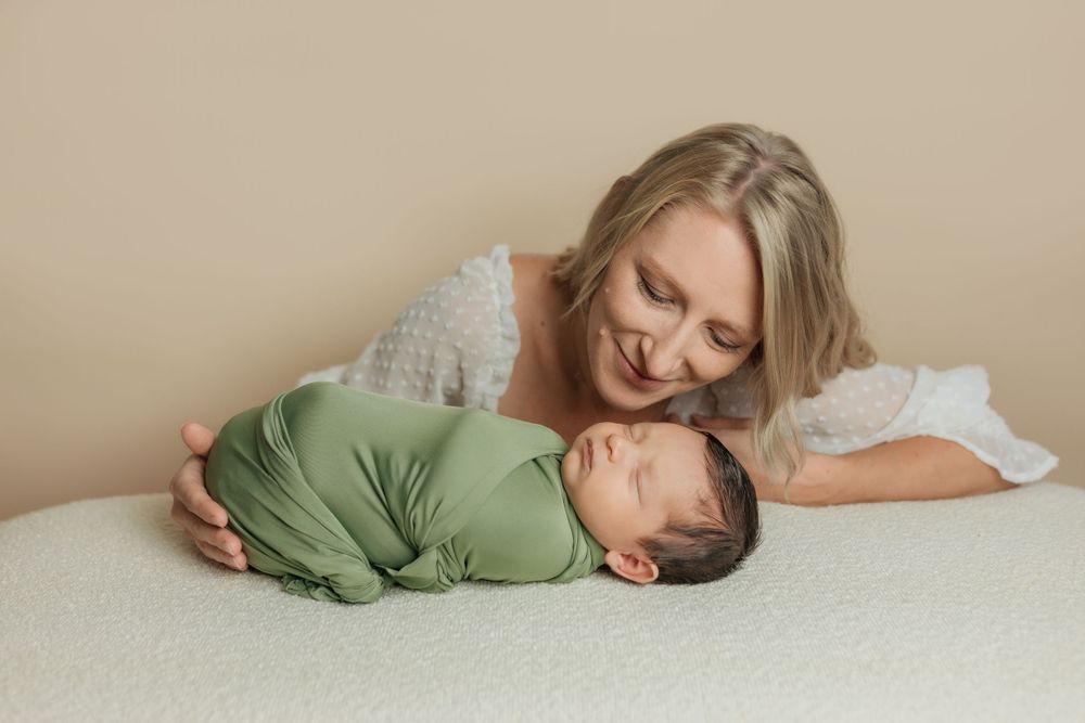 A tender moment captured on white bedding as a mother gently embraces her sleeping newborn wearing green.
