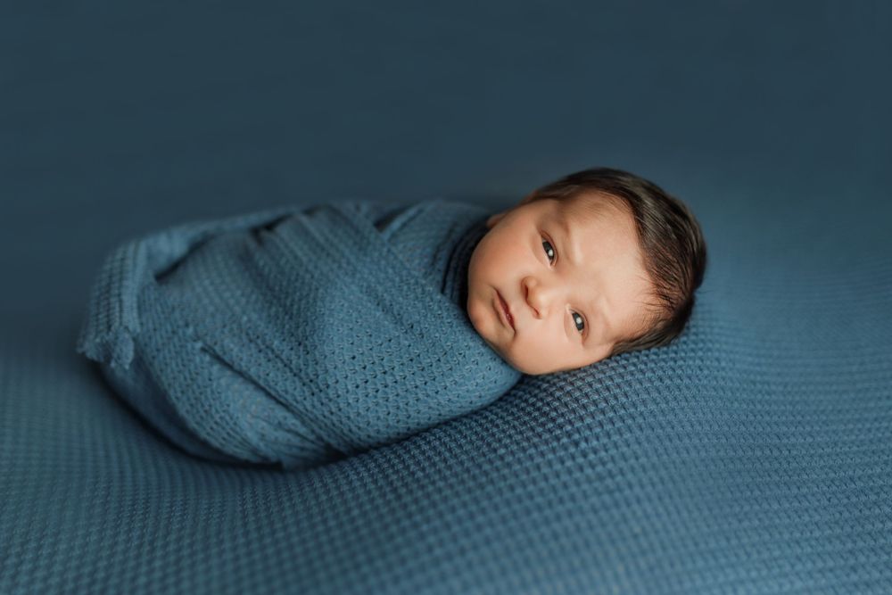 Sleeping newborn baby wrapped in cozy blue swaddle blanket on textured blue background.