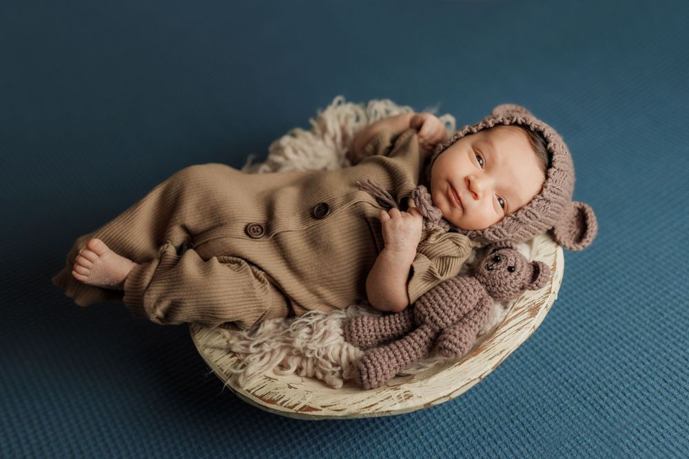 Sleeping newborn wearing brown knit outfit lies peacefully in woven basket on blue backdrop during professional photoshoot.