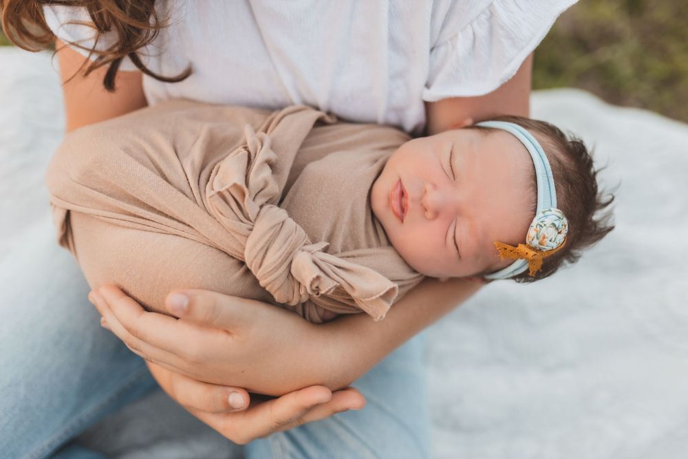 A sleeping newborn baby in a beige swaddle wrap wearing a mint headband rests peacefully in gentle arms.