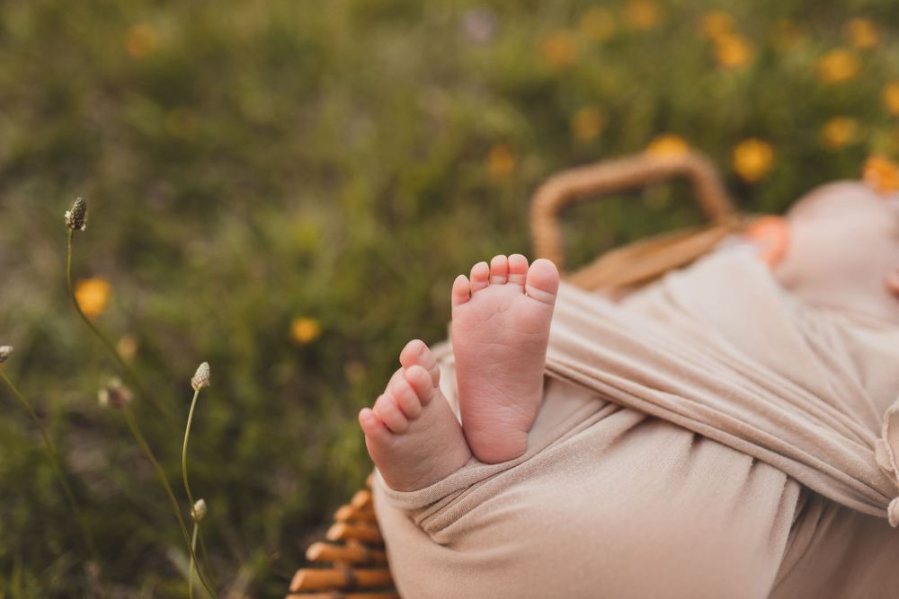 Tiny baby feet resting on beige fabric in a wicker basket outdoors among wildflowers.