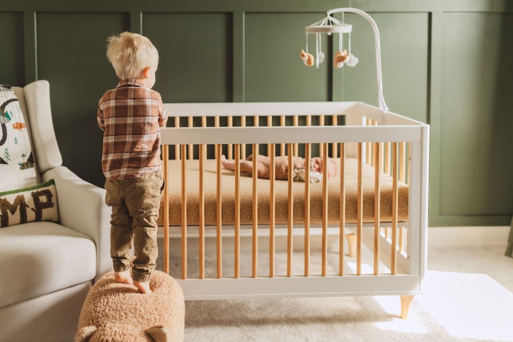 Small child stands on pouf looking into wooden crib with mobile in modern nursery with sage green walls.