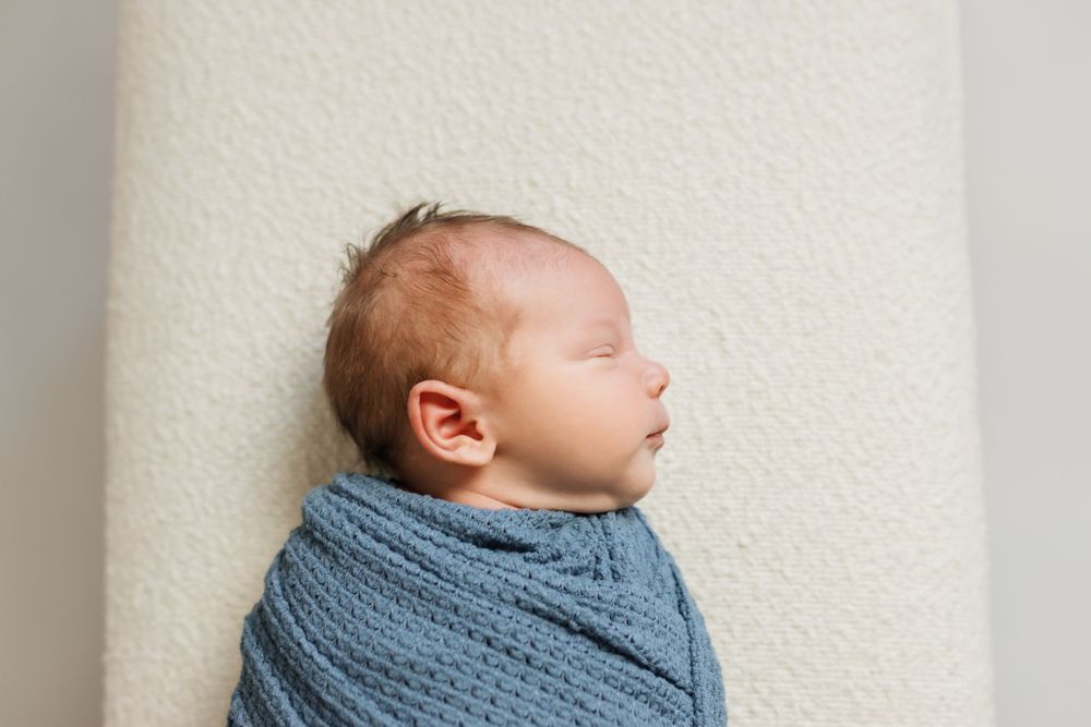 A sleeping newborn baby wrapped in a soft blue knit blanket lies against a cream-colored background.