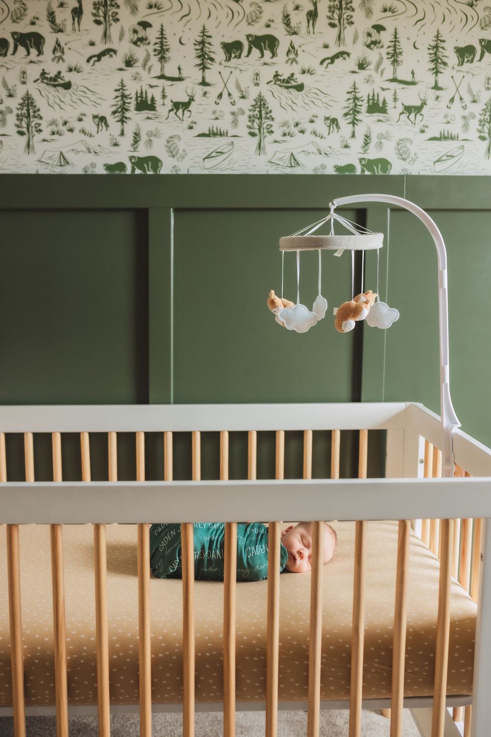 Modern nursery with wooden crib, forest animal wallpaper, and hanging mobile against sage green walls.