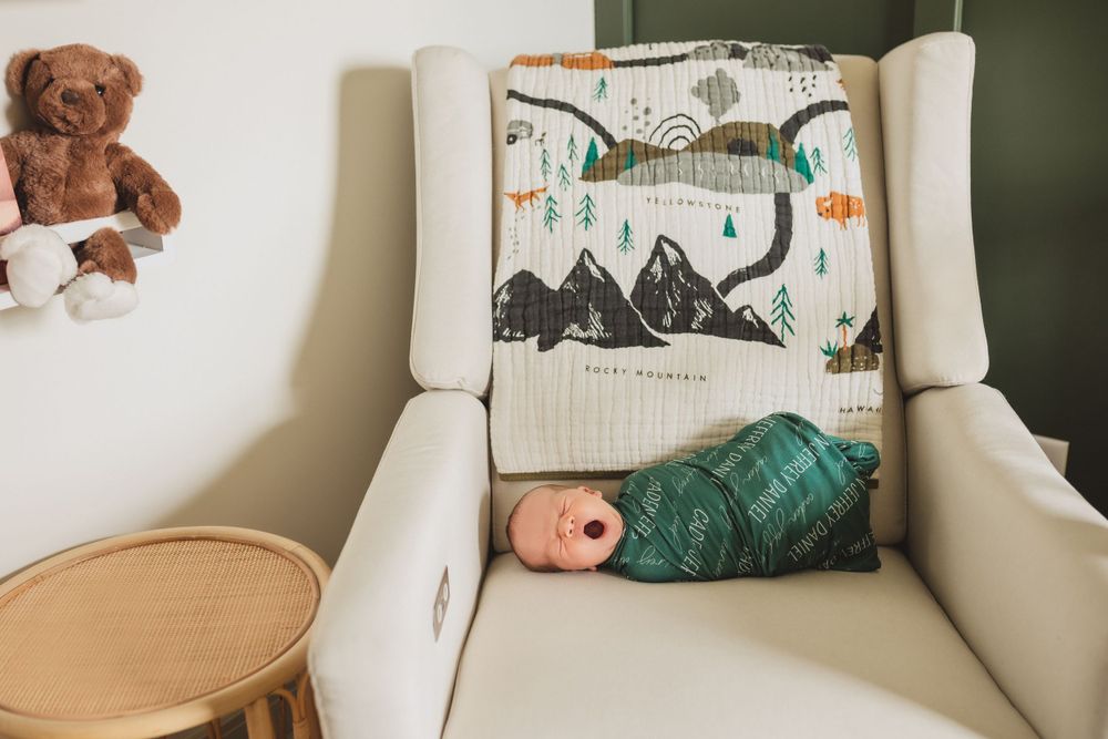Ivory nursery chair with mountain scene pillow, teddy bear and wooden table in cozy corner.
