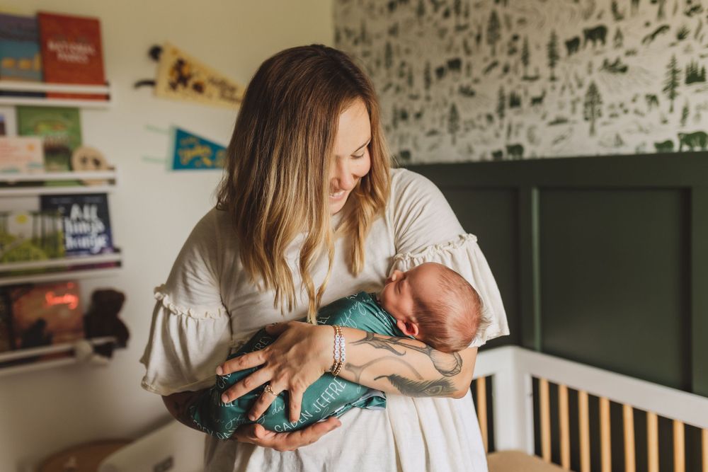 Parent in white robe holding and gazing lovingly at newborn baby in nursery with dark wallpaper and crib.