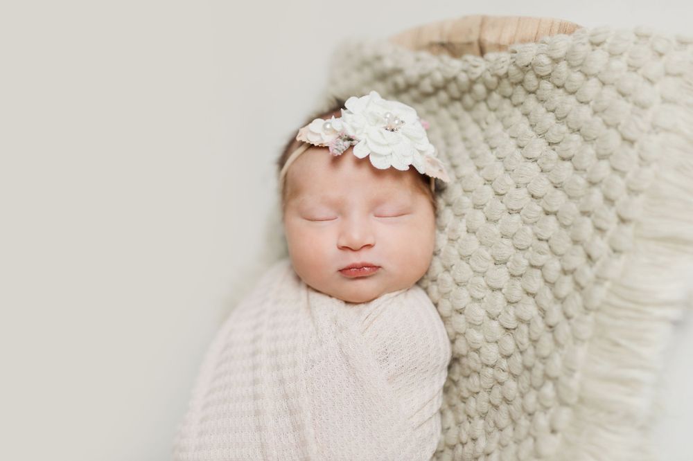 Peaceful sleeping newborn wrapped in white knit blanket laying on textured beige surface with delicate lace headband.