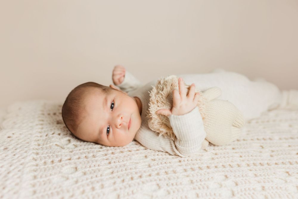 A newborn baby lies on a white knitted blanket wearing a cozy cream-colored outfit while looking at the camera.