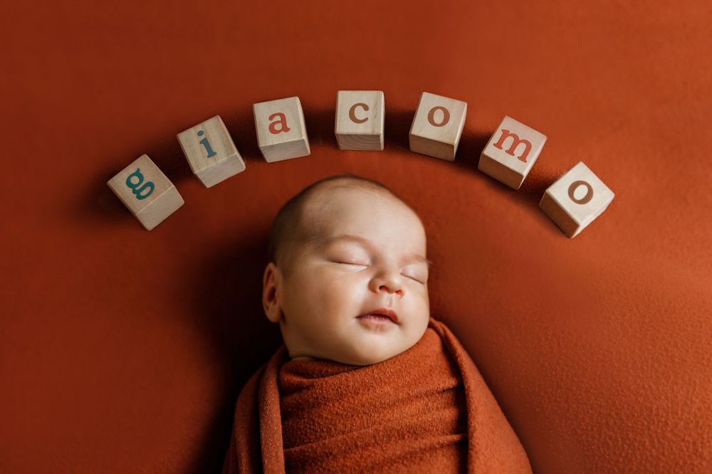 A sleeping newborn swaddled in orange fabric with letter blocks spelling 'welcome' arranged above.