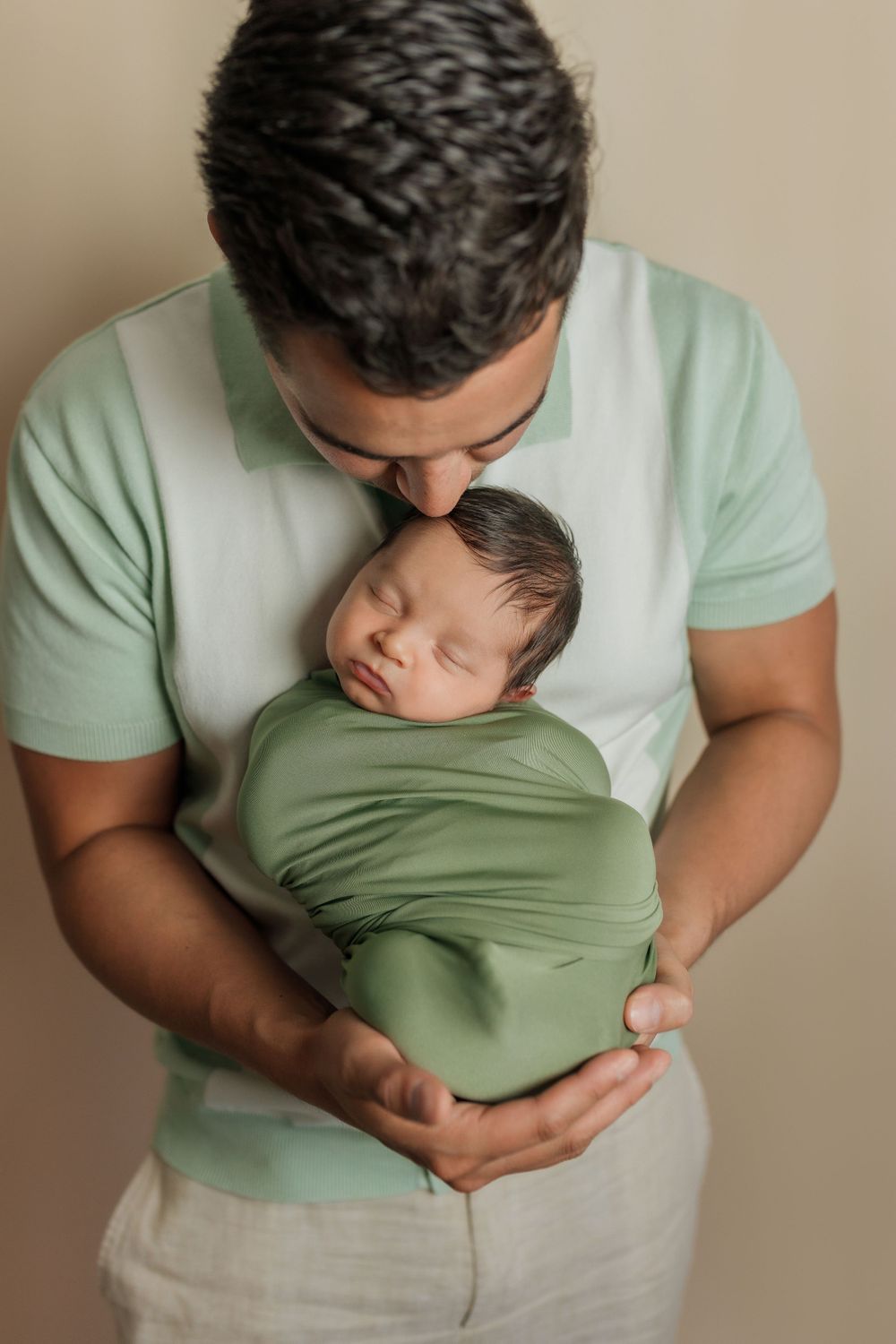 A tender moment between parent and newborn baby wrapped in soft sage green fabric against a neutral backdrop.