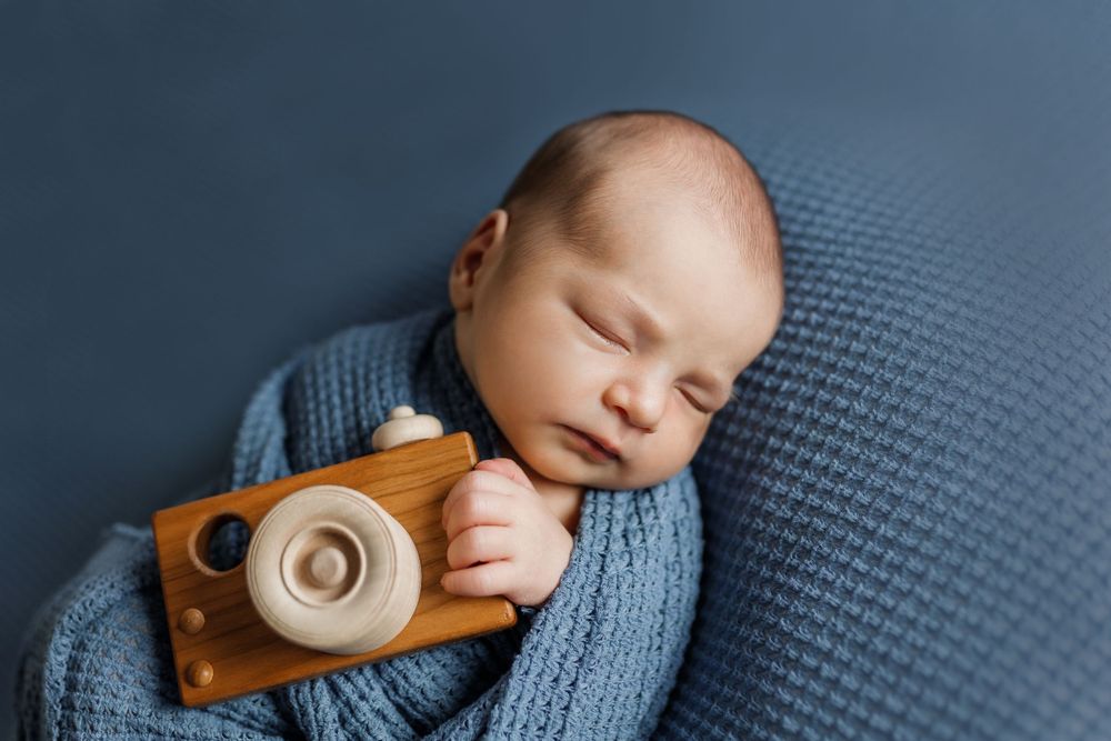 Sleeping newborn wrapped in blue knit blanket holding wooden toy camera against navy background.